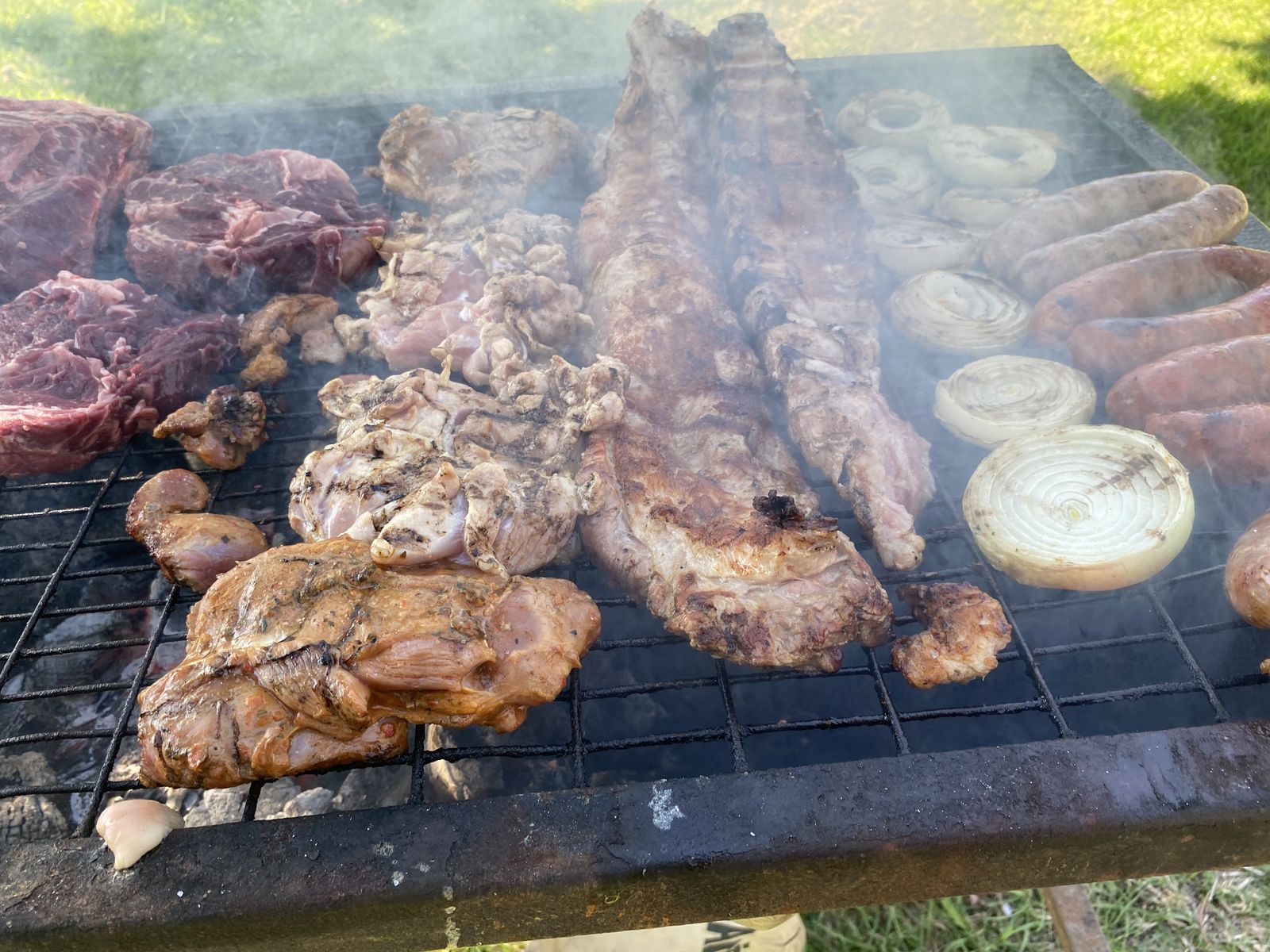 Parrilla al aire libre con carne, longanizas y cebolla, humeando sobre el carbón.